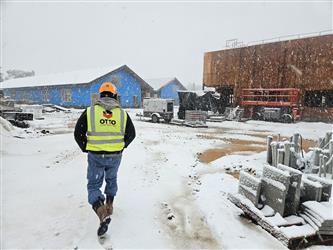 a man walking in a yellow "Otto Construction" high visibility vest in the snow toward two buildings under construction