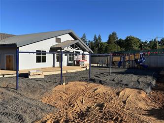 The blue frame of a swing set is set in dirt and gravel in front of a classroom building with more play equipment in the background